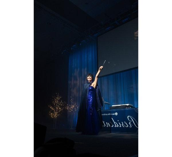 President Philomena Mantella smiles at the crowd while holding a magic wand above her head, and wearing a dark blue cloak over her dress.